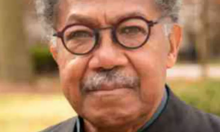 Older man with gray hair and glasses, standing outdoors with a blurred background of trees and grass