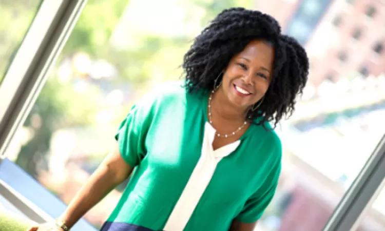 Smiling woman with curly hair wearing a green and white top, standing by a window with city buildings in the background.