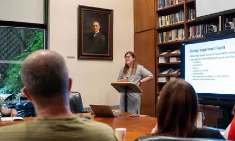 A woman presents at a podium in a library with a portrait and bookshelves. Attendees sit listening.
