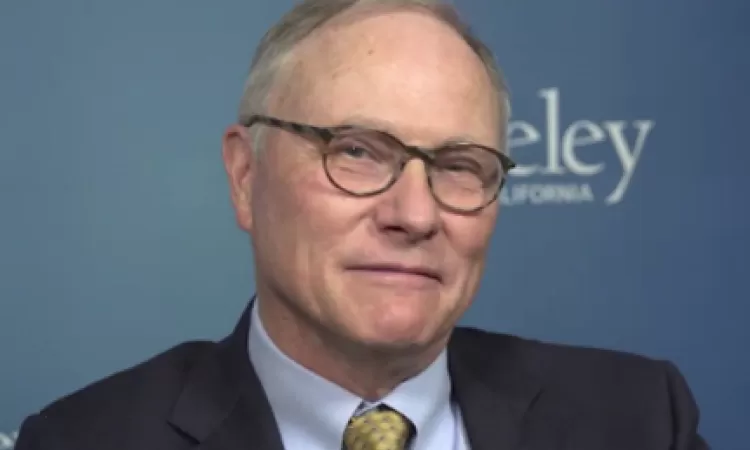 Smiling older man wearing glasses, a suit, and tie, in front of a blue background with the word "Berkeley" partially visible.
