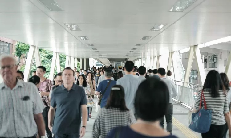 Crowded pedestrian overpass with people walking in both directions, many in casual or business attire, under a covered walkway in an urban setting.