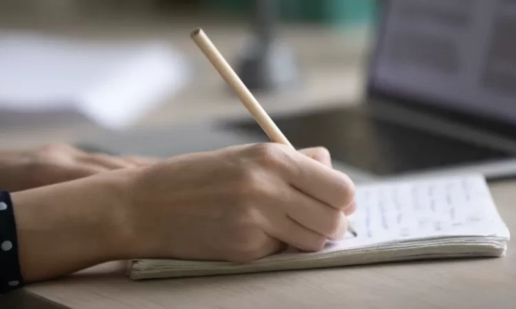 A person writes in a notebook with a pencil, next to an open laptop on a wooden desk.