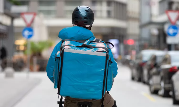 Food delivery cyclist wearing a blue jacket and large blue insulated backpack, riding down a city street with cars parked along the side.