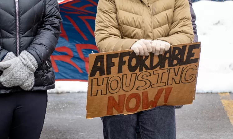 Person wearing a thick coat and gloves outside with a cardboard sign that says "Affordable Housing Now!"