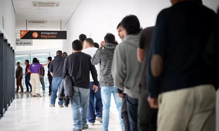 Several people waiting in a line indoors under a sign that reads "Immigration - Migración"