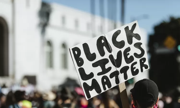Person holding a “Black Lives Matter” sign at a protest, with a blurred crowd and building in the background.